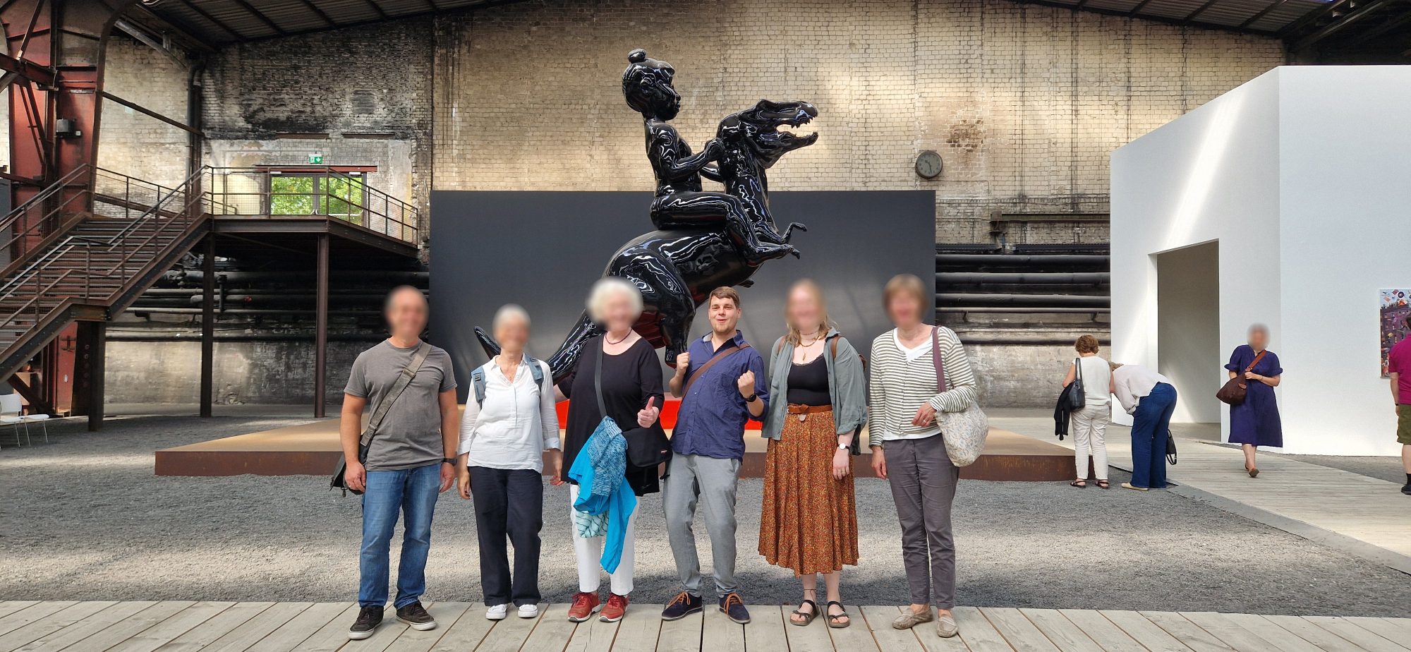 Kursgruppe bei einer Ausstellungs-Exkursion vor einer großen schwarzen Skulptur in einer Industriehalle